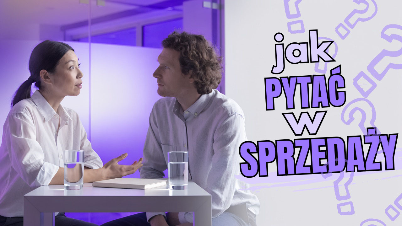 Dwie osoby rozmawiają przy stole w biurze, jedna zadaje pytanie, druga odpowiada.